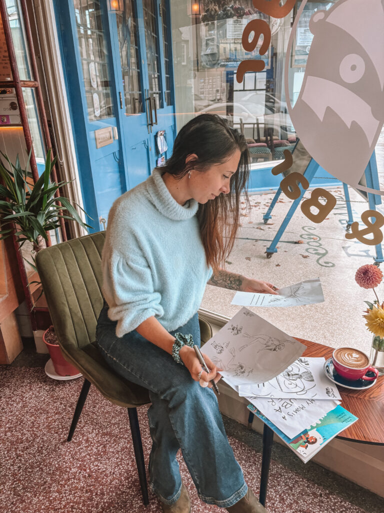 Sarah sits at a table in the window of a coffee shop. She is looking over draft pages of illustrations. 